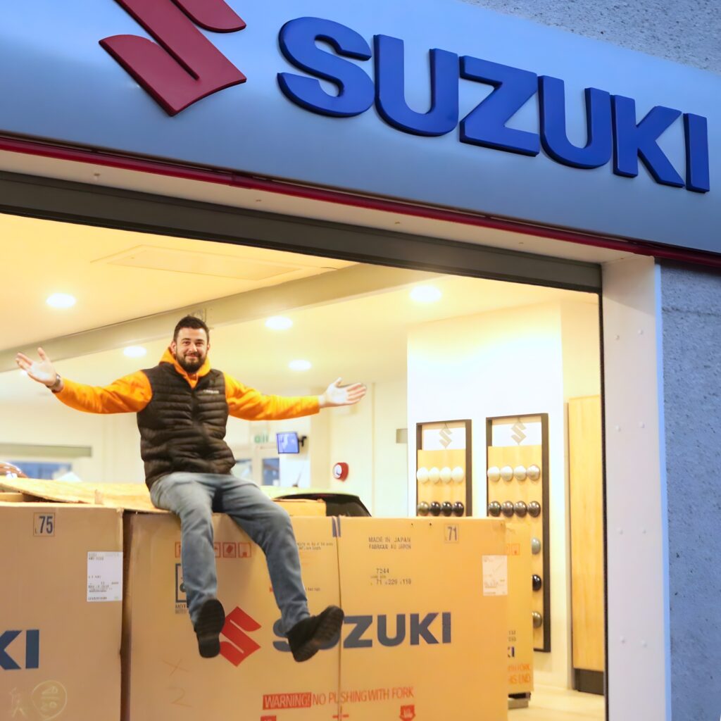 Image of Thomas with Suzuki sign sitting on a Suzuki box that has a bike inside.